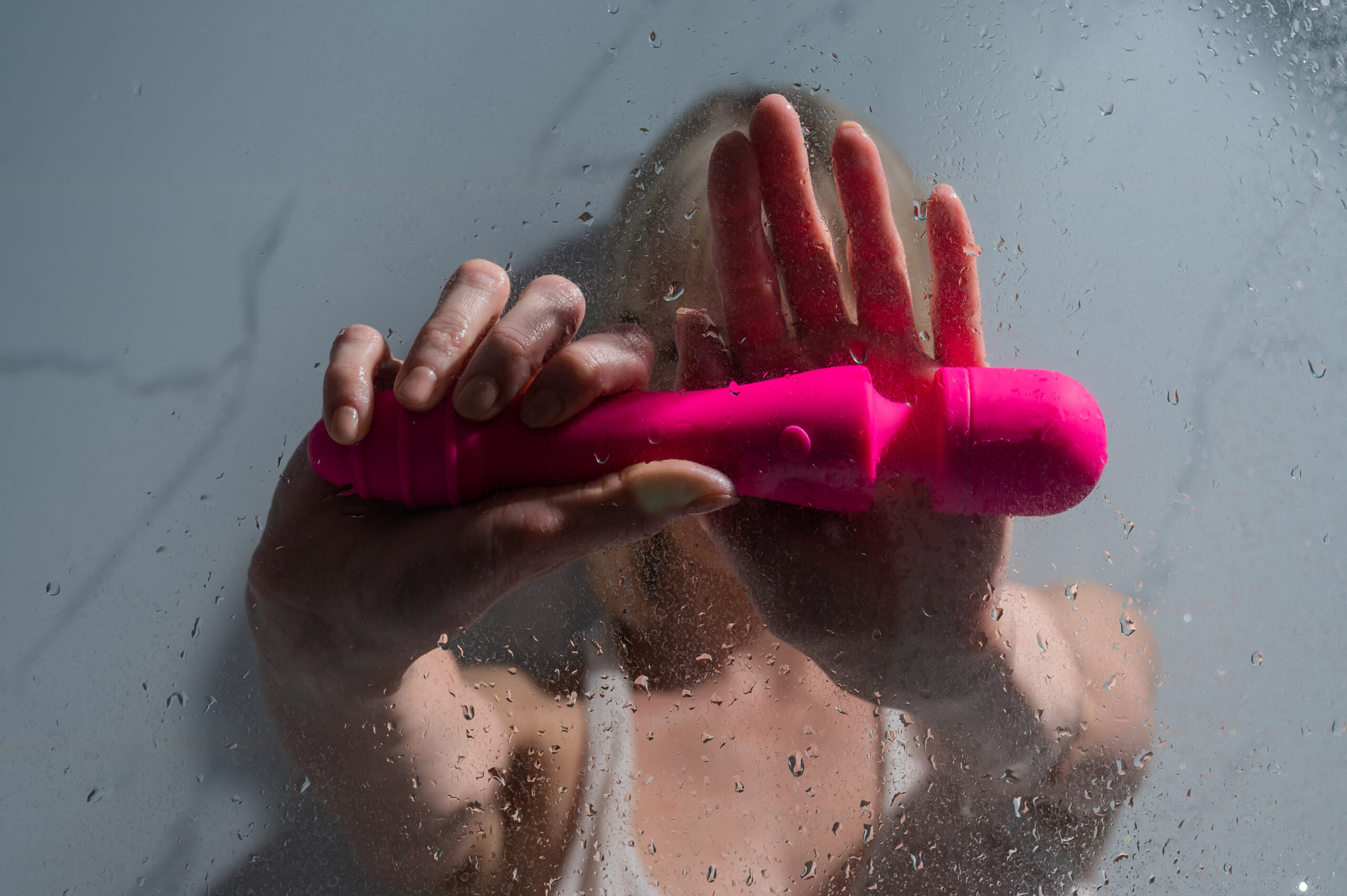 A woman in a white tank top stands in the shower and holds a pink sex toy.